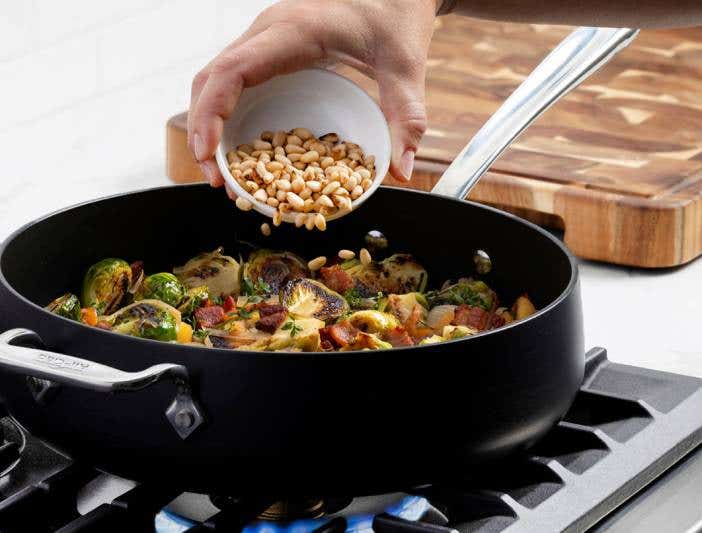 A person stirring beans in an All-Clad HA1 Expert Nonstick pan on a stove, showcasing cooking in action.
