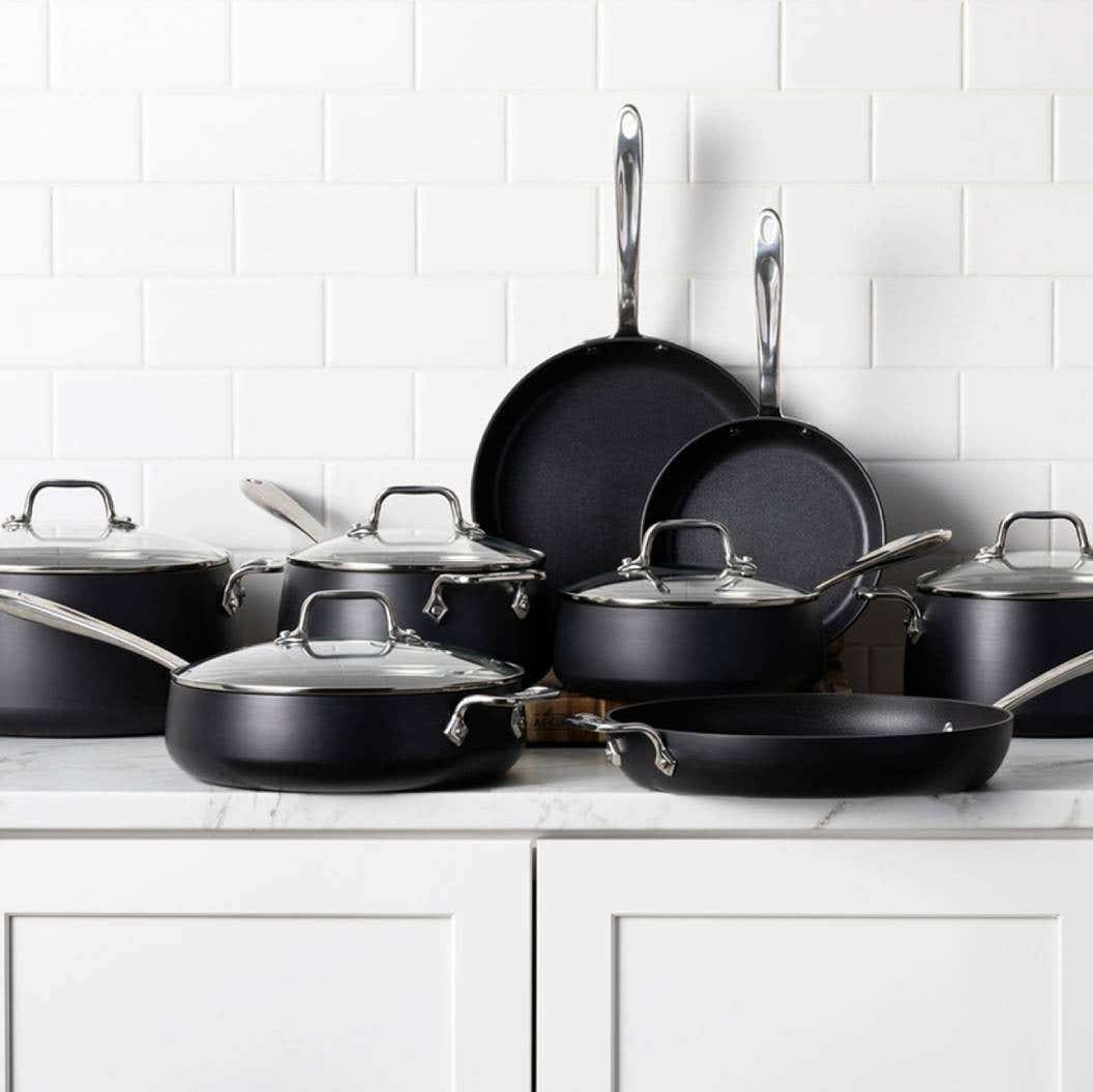 A collection of black All-Clad HA1 Expert nonstick pots and pans arranged neatly on a kitchen counter.
